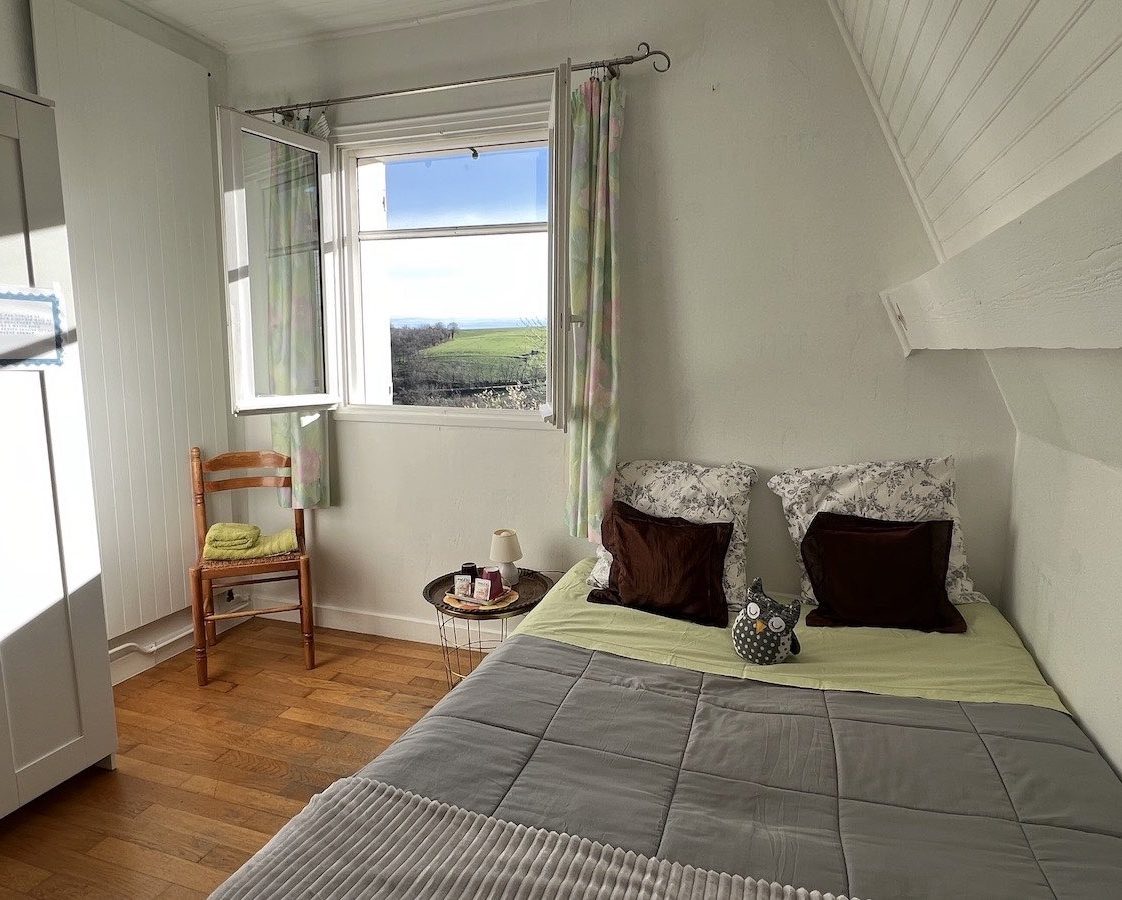 Vue lit enfants couverture grise, fenêtre ouverte avec moustiquaire, chaises et serviettes de toilette vertes, plateau d'accueil