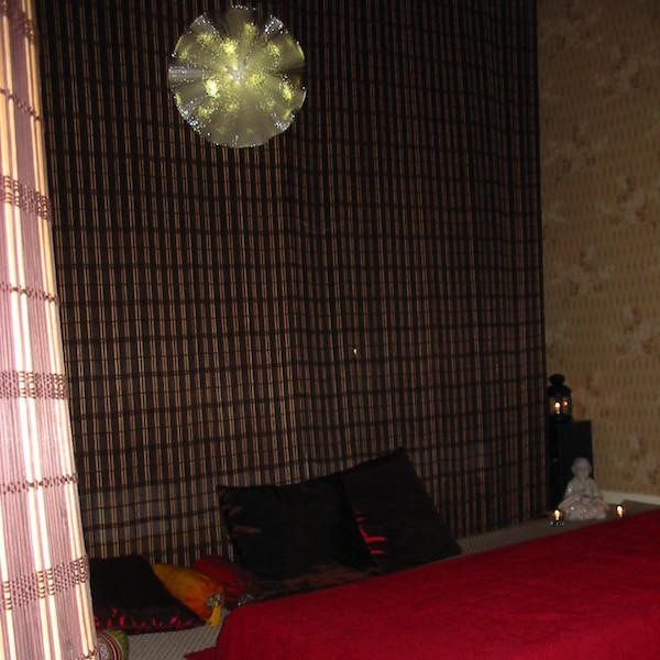 Salle de massage avec table et coussins rouge, rideaux de bambous marrons et lumière au plafond.