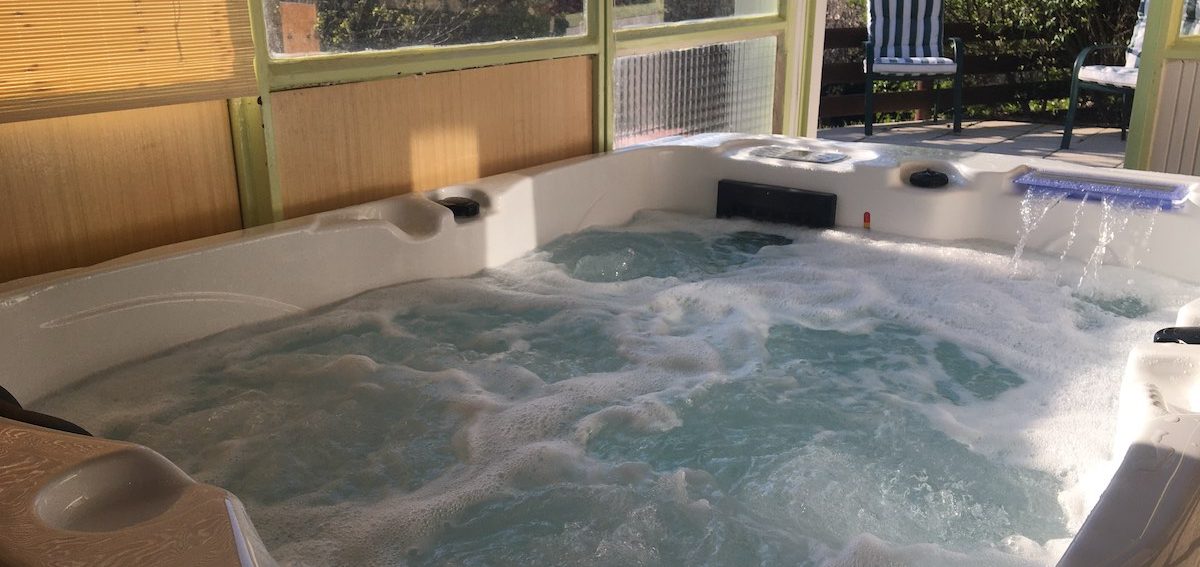 Jacuzzi privatif en véranda nature, porte ouverte sur la terrasse avec vue sur 2 fauteuils confortables 