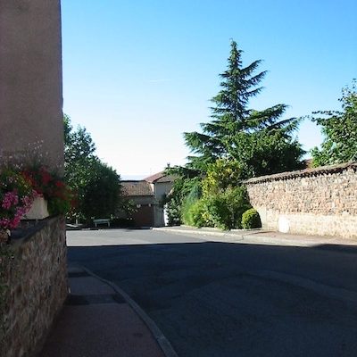 Vue de la rue principale verdoyante de Maringes en direction du stade, arbres à droite, fleurs à gauche