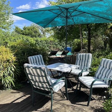Terrasse avec quatre fauteuils et petite table, parasol bleu et aperçu piscine dans le jardin en fond.