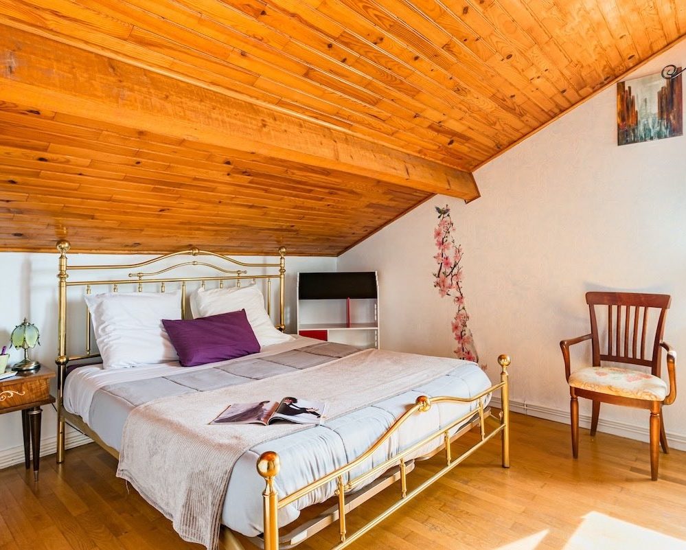 Chambre iris avec grand lit queen, table de chevet avec lampe verte, petit fauteuil, plafond mansardé en bois naturel et plancher.